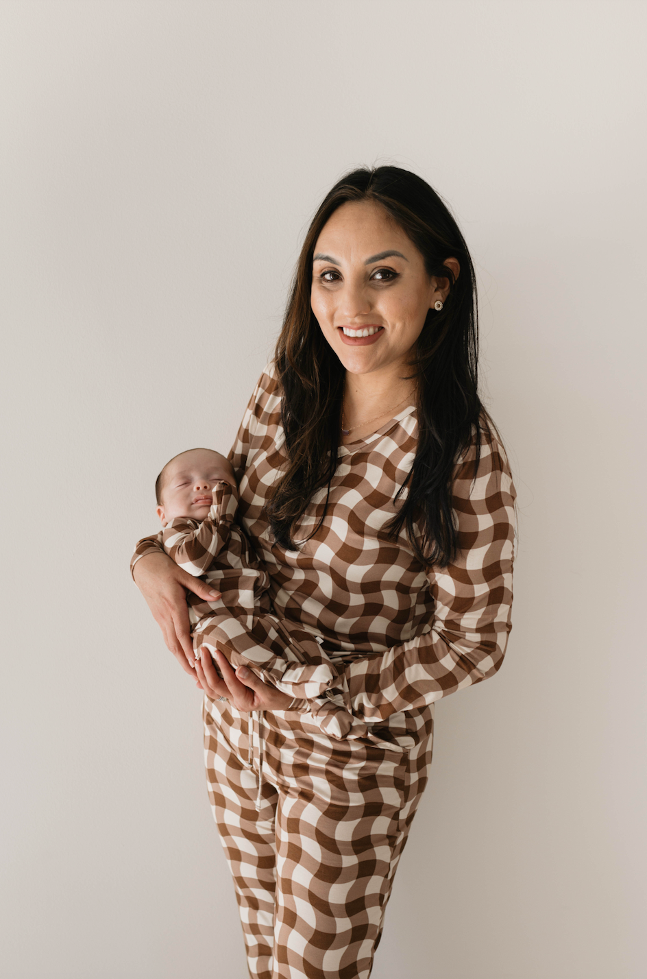 A woman wearing Women's Bamboo Pajamas in Groovy Gingham by forever french baby holds a sleeping baby against a plain white background. Both have dark hair. The woman, likely one of the many breastfeeding moms who appreciate bamboo fabric's softness, is smiling and looking at the camera.