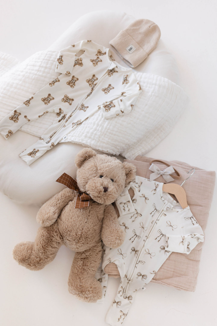 Flat lay of baby items including a white onesie with a teddy bear print, a beige knit hat, Kendy Du's Bamboo Two Piece Pajamas | Clara Bow, a plush teddy bear with a brown ribbon, a white quilted blanket, and a beige swaddle. The setting is a clean, white background.