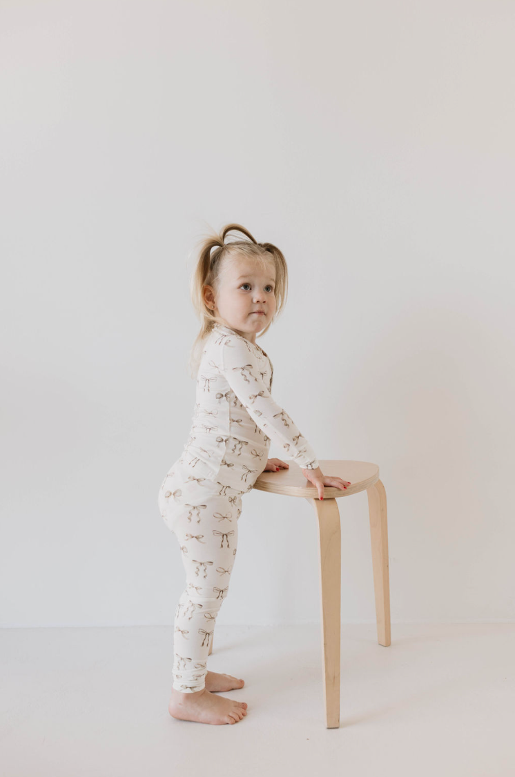 A young child with blond hair in pigtails stands barefoot beside a wooden stool. The child is dressed in Kendy Du's hypo-allergenic Bamboo Two Piece Pajamas | Clara Bow, featuring light brown animal prints. With both hands resting on the stool, the child gazes slightly off to the side against a plain white background.