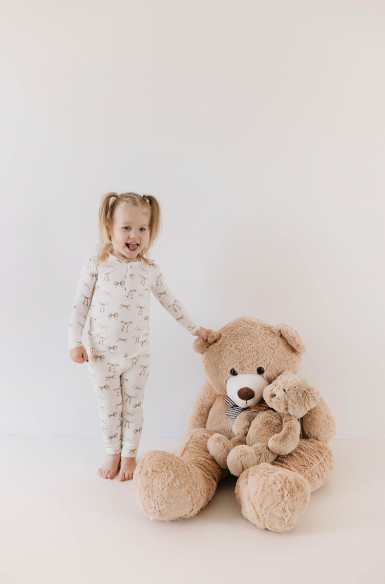 A young child with blonde pigtails stands barefoot in her Kendy Du Bamboo Two Piece Pajamas | Clara Bow, holding the paw of an oversized stuffed teddy bear. A smaller teddy bear sits on the larger bear's lap. The background is plain white, creating a clean and simple setting.