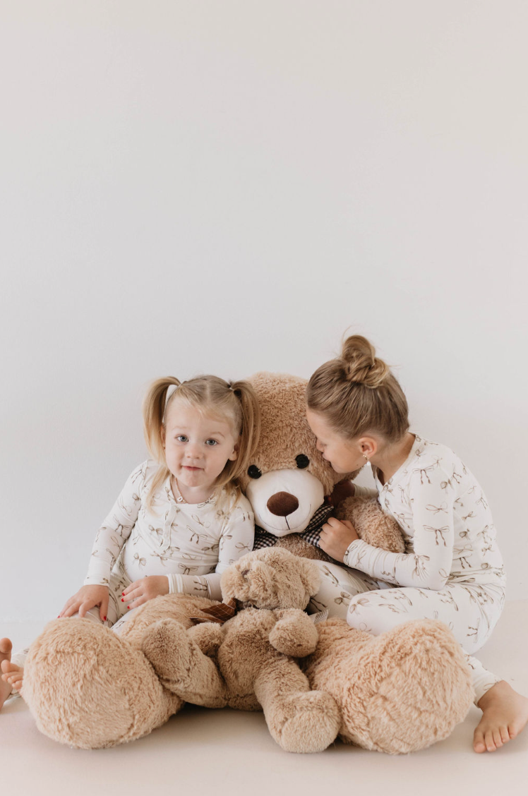 Two young children in matching Kendy Du Bamboo Two Piece Pajamas | Clara Bow with animal prints sit on the ground, embracing a large teddy bear. One child looks at the camera while the other leans into the stuffed bear, holding a smaller teddy bear. The background is plain light-colored, showcasing their hypo-allergenic sleepwear from Forever French Baby.