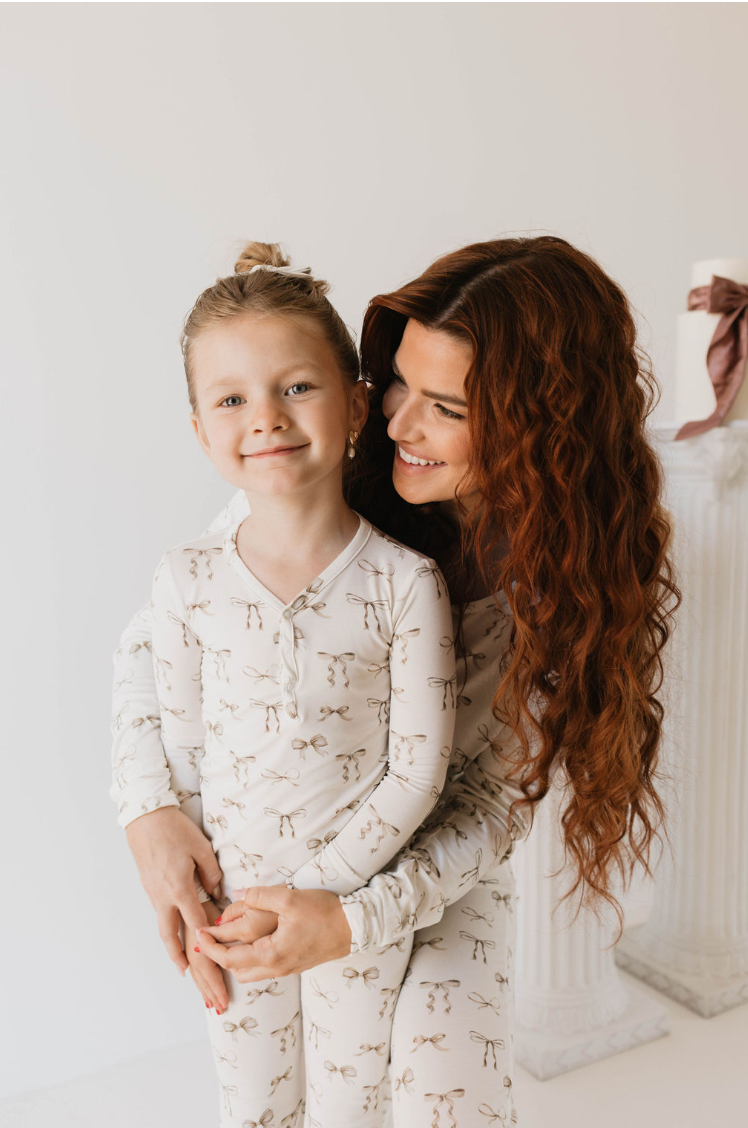 A woman with long curly hair embraces a smiling young girl with a top bun. Both are wearing matching white Bamboo Two Piece Pajamas, Clara Bow from the Kendy Du brand, which feature a delicate floral pattern. The background showcases a white column with a ribbon, set against a pristine white wall.