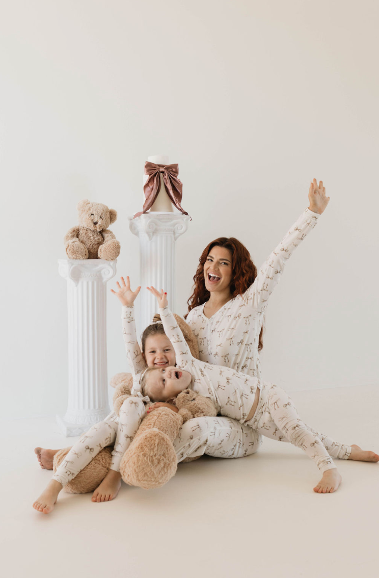 A woman and two children dressed in matching, hypo-allergenic Bamboo Two Piece Pajamas | Clara Bow by Kendy Du, featuring a charming teddy bear pattern, pose joyfully in front of white pillars. The children lay on a large teddy bear on the floor, while the woman sits behind them, all raising their arms in celebration.