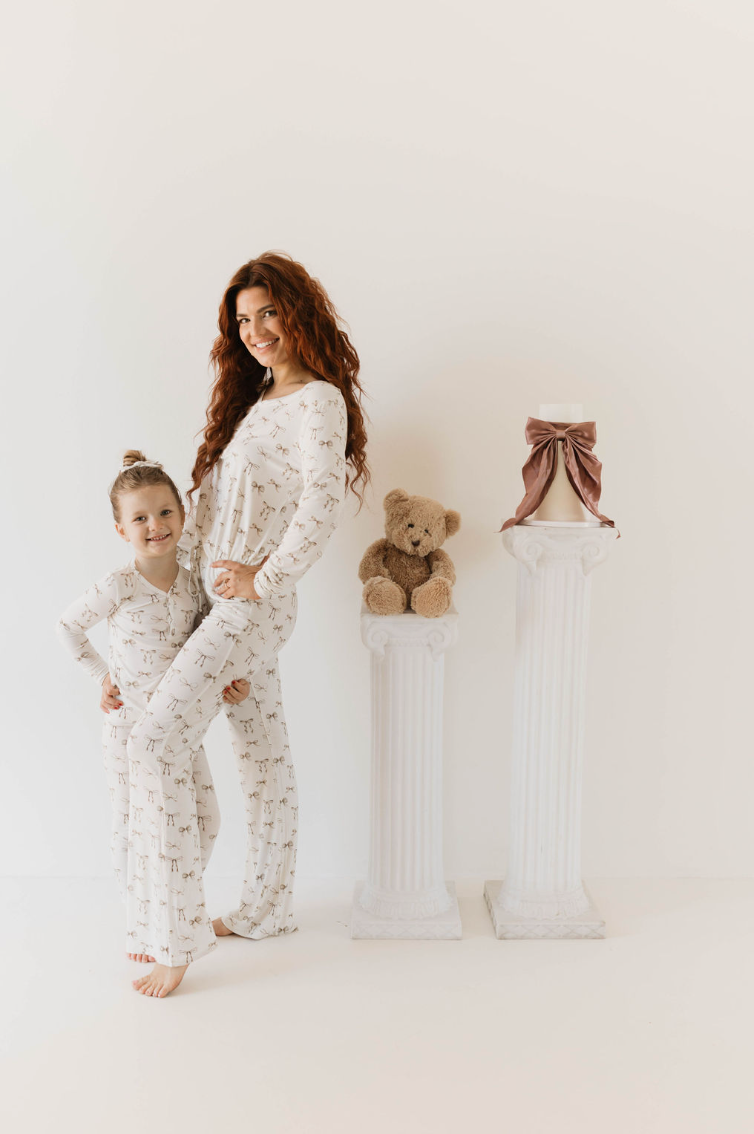 A woman and a young girl, both with long hair, are wearing matching white Kendy Du Bamboo Two Piece Pajamas | Clara Bow with small prints. They are posing side by side and smiling brightly. Behind them, there are two white pillars, one with a teddy bear and the other with a cake adorned with a pink ribbon.