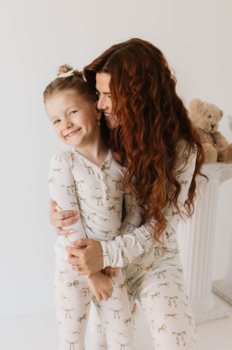 A woman with long, curly red hair hugs a smiling young girl with blonde hair. Both are wearing matching Kendy Du Bamboo Two Piece Pajamas | Clara Bow made from hypo-allergenic sleepwear. They are standing in a bright room with a white pedestal and a beige teddy bear in the background.