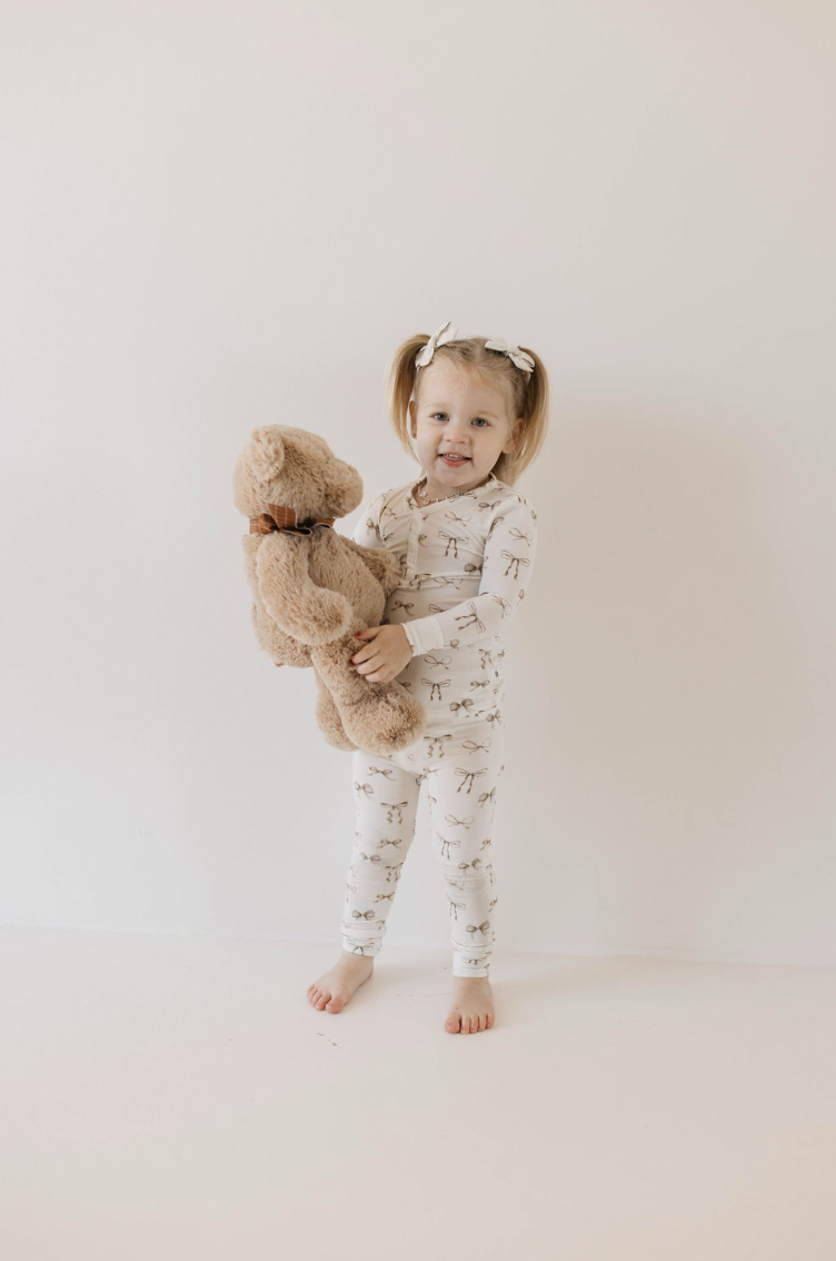 A young child with pigtails, dressed in a white Kendy Du Bamboo Two Piece Pajamas - Clara Bow set with a pattern, stands barefoot against a plain background, holding a large plush teddy bear. The child is smiling slightly, and the teddy bear is wearing a brown bow around its neck.