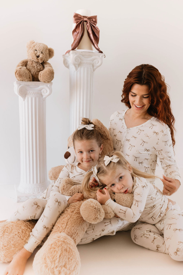 A woman and two young girls, all in matching Bamboo Two Piece Pajamas | Clara Bow from Kendy Du, pose with a giant teddy bear. The girls hug the teddy bear while the woman sits behind them with a smile. Two white columns in the background hold another teddy bear and a large candle.