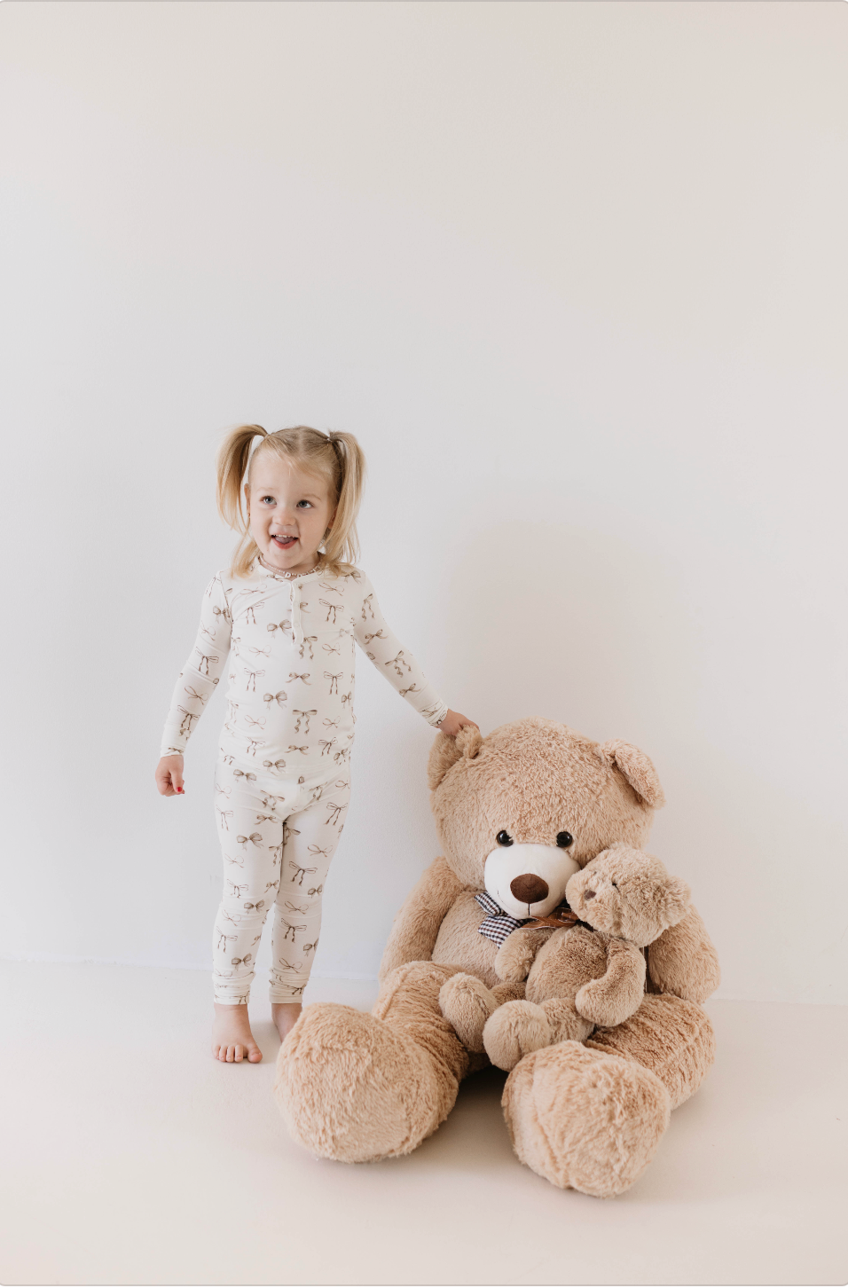 A young child with blonde pigtails stands barefoot next to a large beige teddy bear and a smaller teddy bear, wearing white Clara Bow Bamboo Two Piece Pajamas by Kendy Du with a giraffe pattern. Smiling and pointing a finger, the child embodies pure joy against a plain white background.