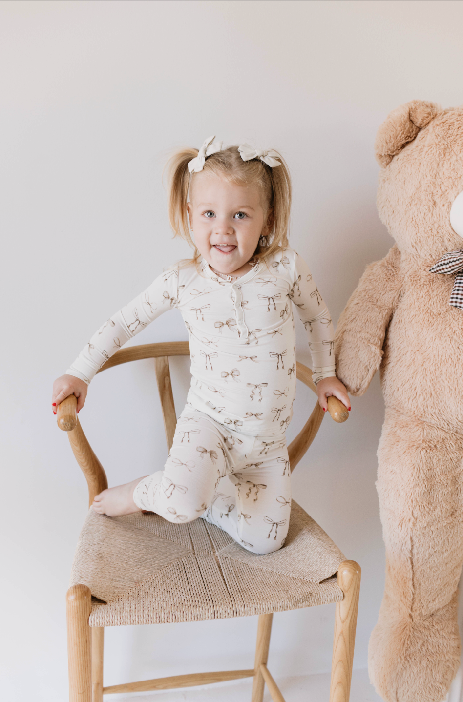 A young child with two pigtails, each tied with white fabric bows, is sitting on a wooden chair. The child is wearing Kendy Du's Bamboo Two Piece Pajamas in the Clara Bow design—matching cream-colored hypo-allergenic sleepwear featuring a light brown pattern—and has one arm around a large teddy bear next to the chair.