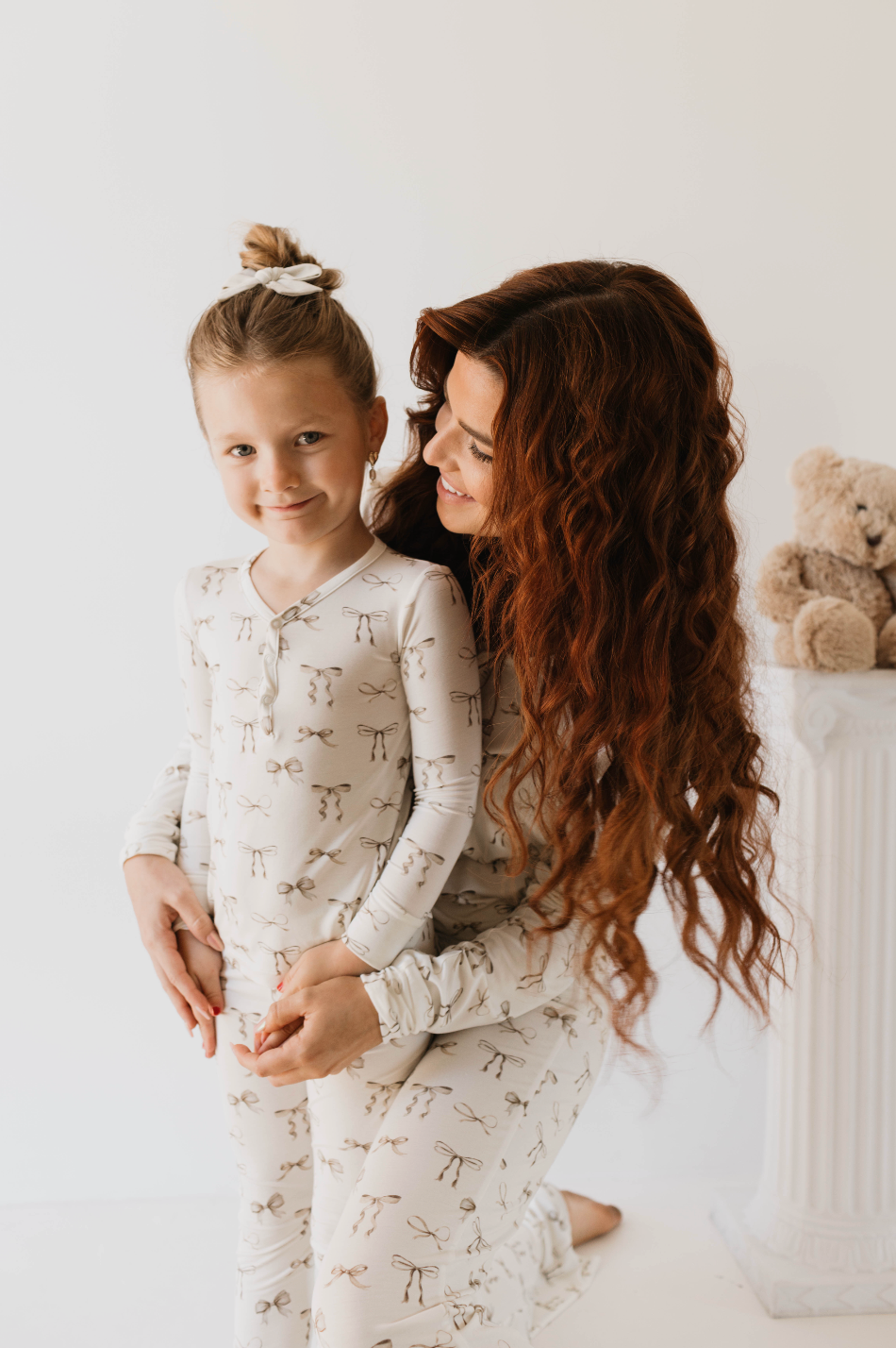 A woman with long, wavy red hair embraces a young girl with blonde hair tied in a bun. Both are smiling and wearing matching light-colored, hypo-allergenic Bamboo Two Piece Pajamas in the Clara Bow design from Kendy Du, featuring animal print patterns. They are standing in front of a white background with a teddy bear on a pedestal.