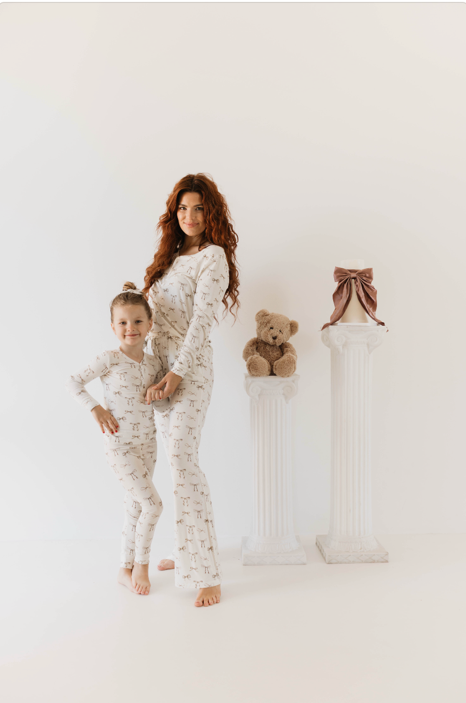 A woman with long, red hair and a young girl stand together in matching hypo-allergenic Bamboo Two Piece Pajamas. The pajamas, from the Kendy Du brand's Clara Bow collection, feature small designs and a crisp white color. They pose next to two white columns; one holds a brown teddy bear, while the other is topped with a pink bow. The background is plain white.