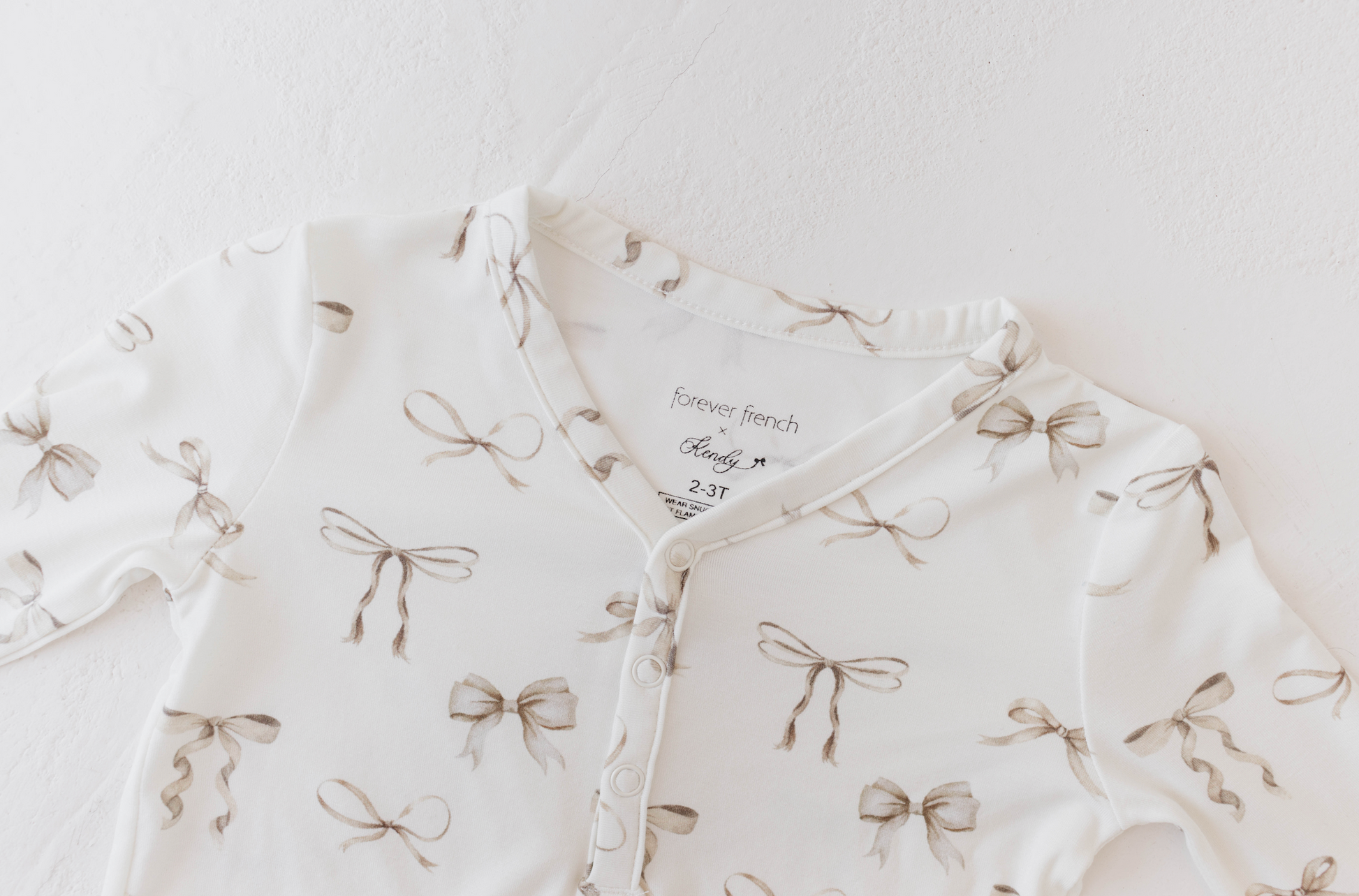 Introducing the Bamboo Two Piece Pajamas | Clara Bow by Kendy Du: a delicate white baby pajama set adorned with a charming pattern of light brown bows. This hypo-allergenic sleepwear features snap buttons, long sleeves, and an inner tag in the collar that reads "Forever French Baby," followed by the size "3-6 months." Crafted with care to ensure ultimate comfort for your little one.