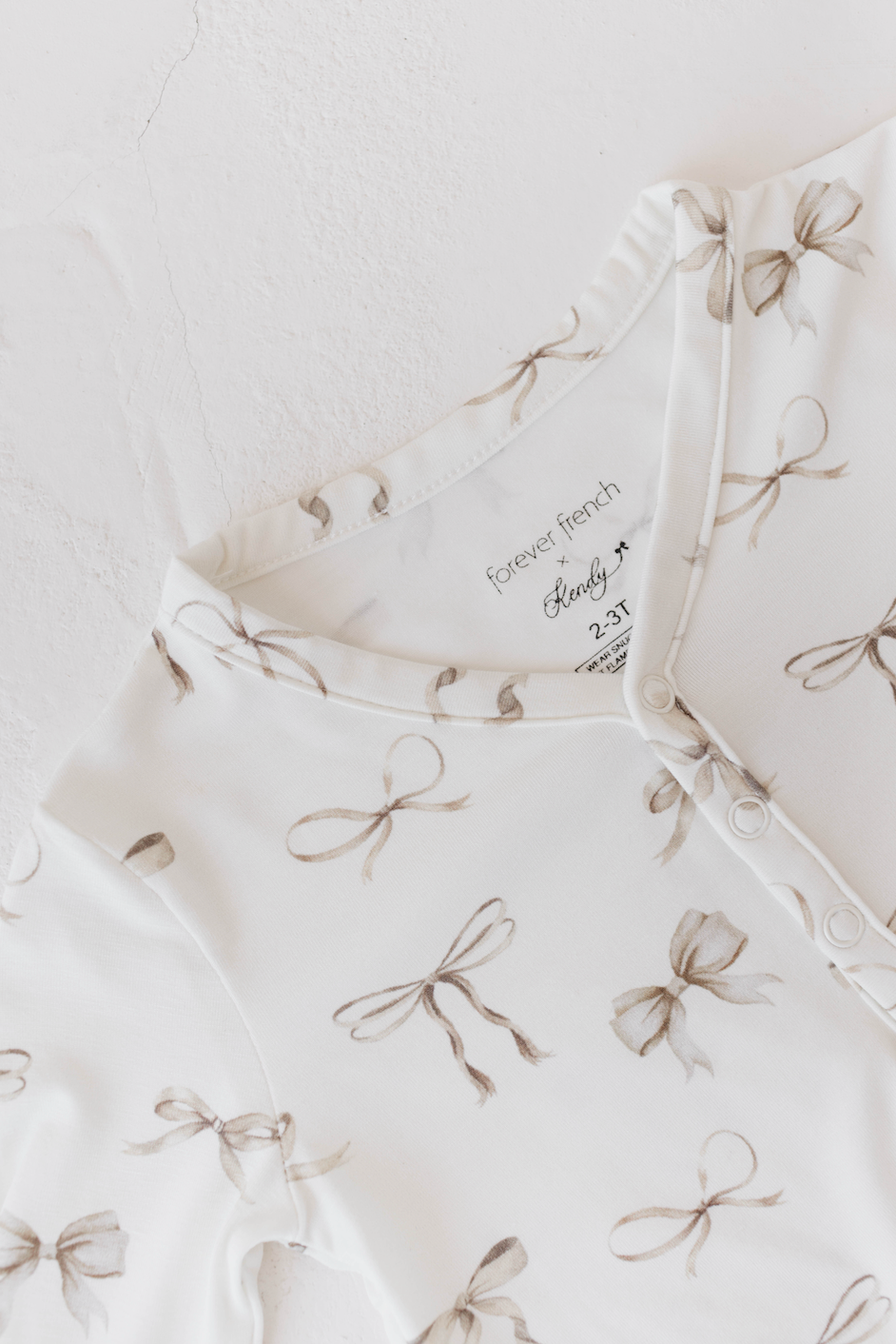 A white baby onesie with a delicate pattern of brown bows, ideal as Kendy Du's hypo-allergenic sleepwear. It features a round neckline with ruffled trim and a button-front closure. The fabric label inside reads "Kendy Du, Clara Bow 2-3T." The onesie is laid out flat on a white surface.