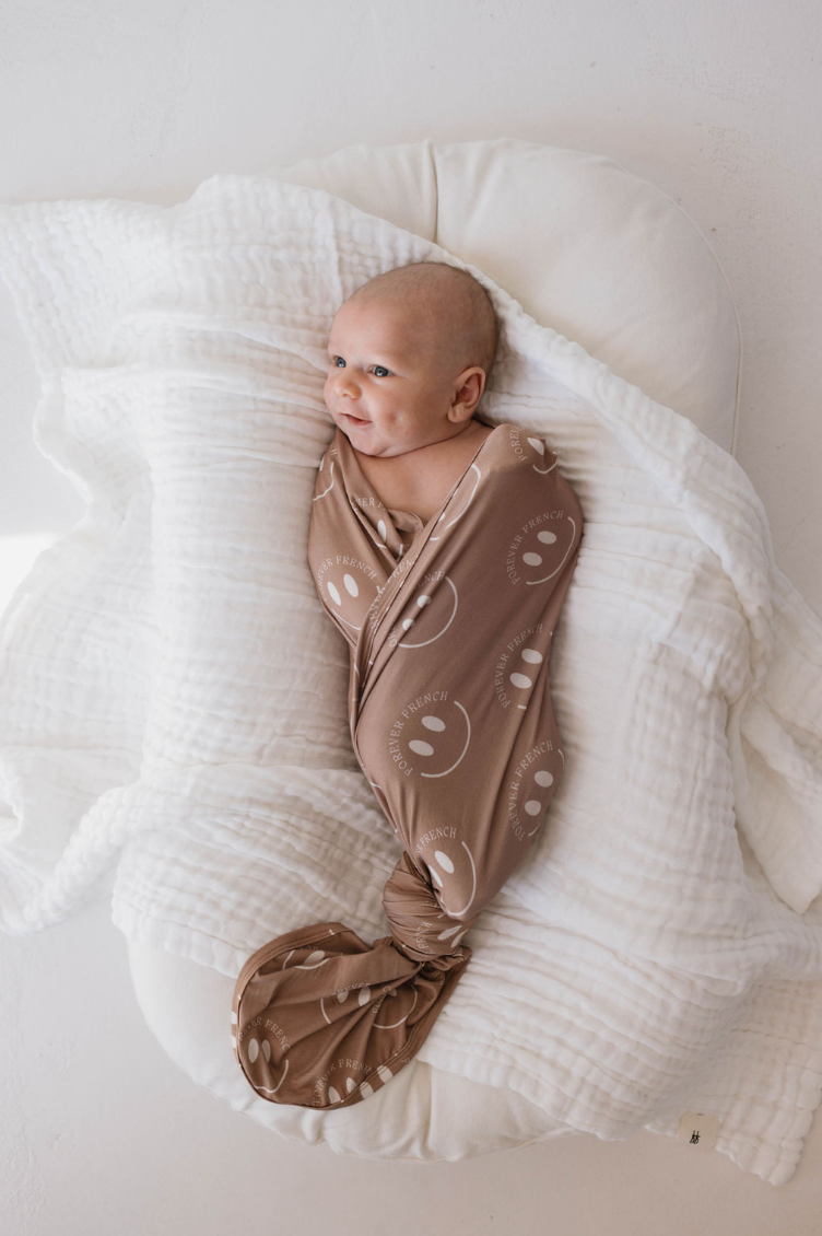 A baby is swaddled in a hypo-allergenic Bamboo Swaddle | Desert Clay ff Smile by forever french baby, lying on a white cushion. The baby is looking to the side with a calm expression. The white cushion and swaddle provide a soft and cozy environment.