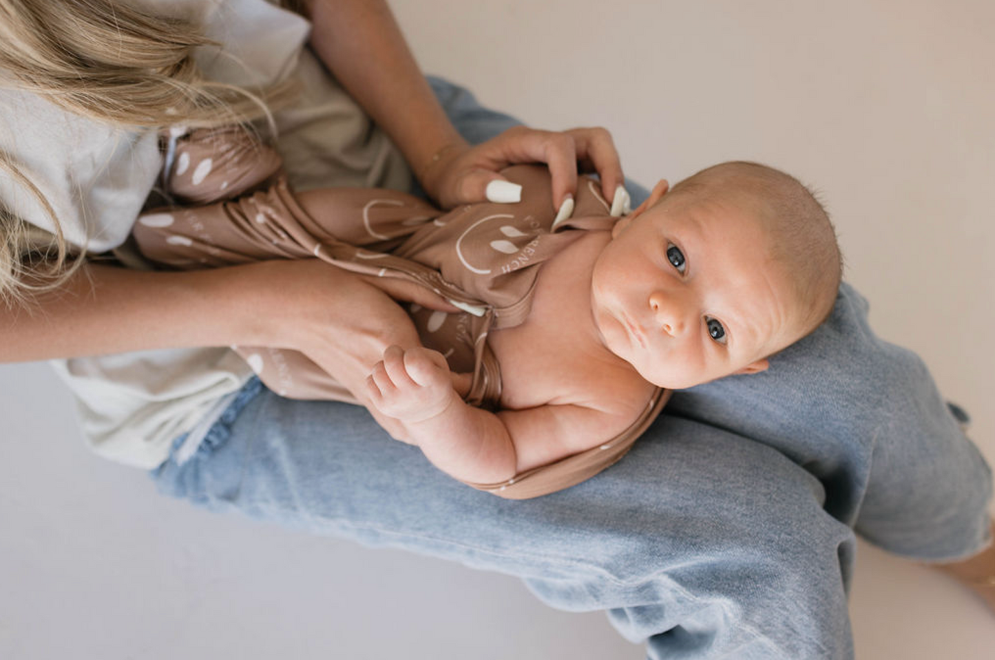 A baby with light-colored hair is swaddled in the Bamboo Swaddle | Desert Clay ff Smile from forever french baby, lying on an adult's lap. The adult, dressed in jeans, is gently cradling the baby with both hands. The baby's wide eyes look curiously ahead.