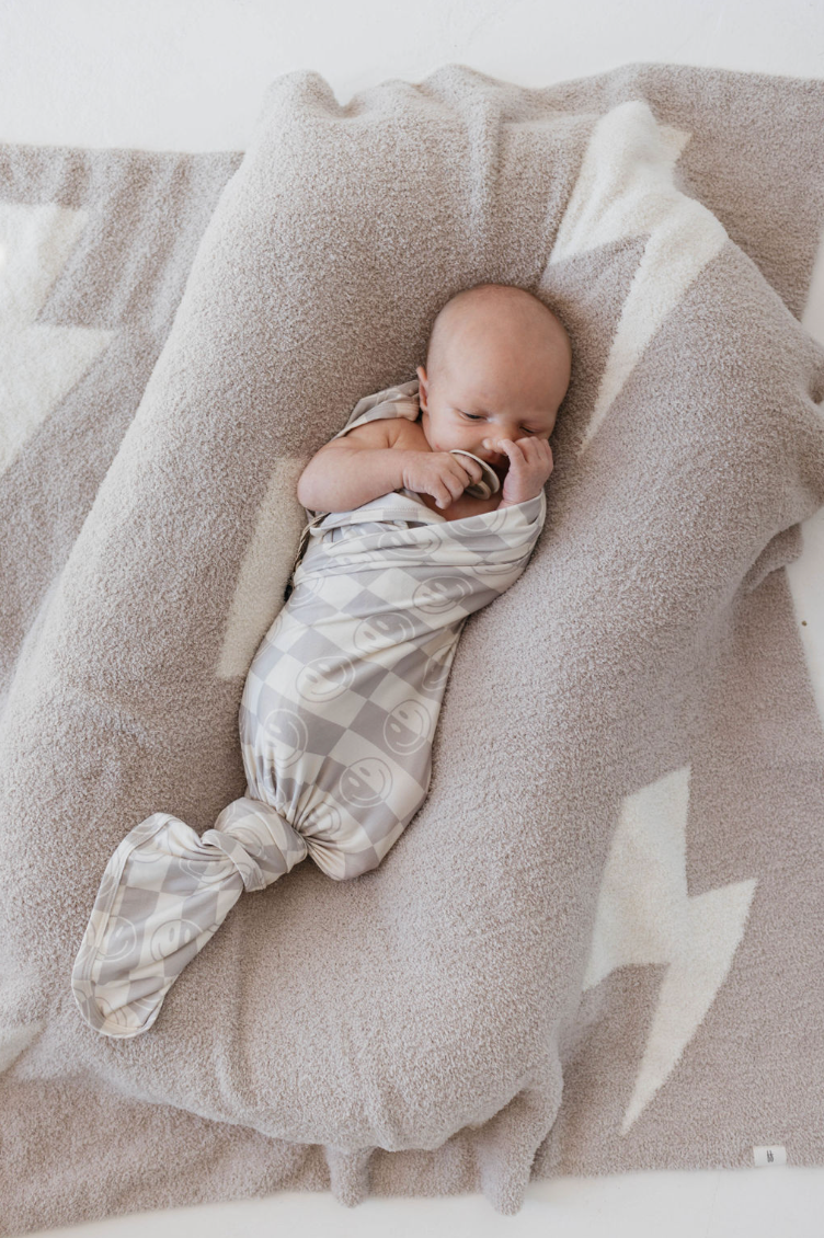 A baby is swaddled in a hypo-allergenic, Smile Checkerboard blanket made of soft bamboo fabric from forever french baby, lying on a plush gray cushion with white zigzag patterns. The baby is sucking on a pacifier and appears to be asleep or resting peacefully.