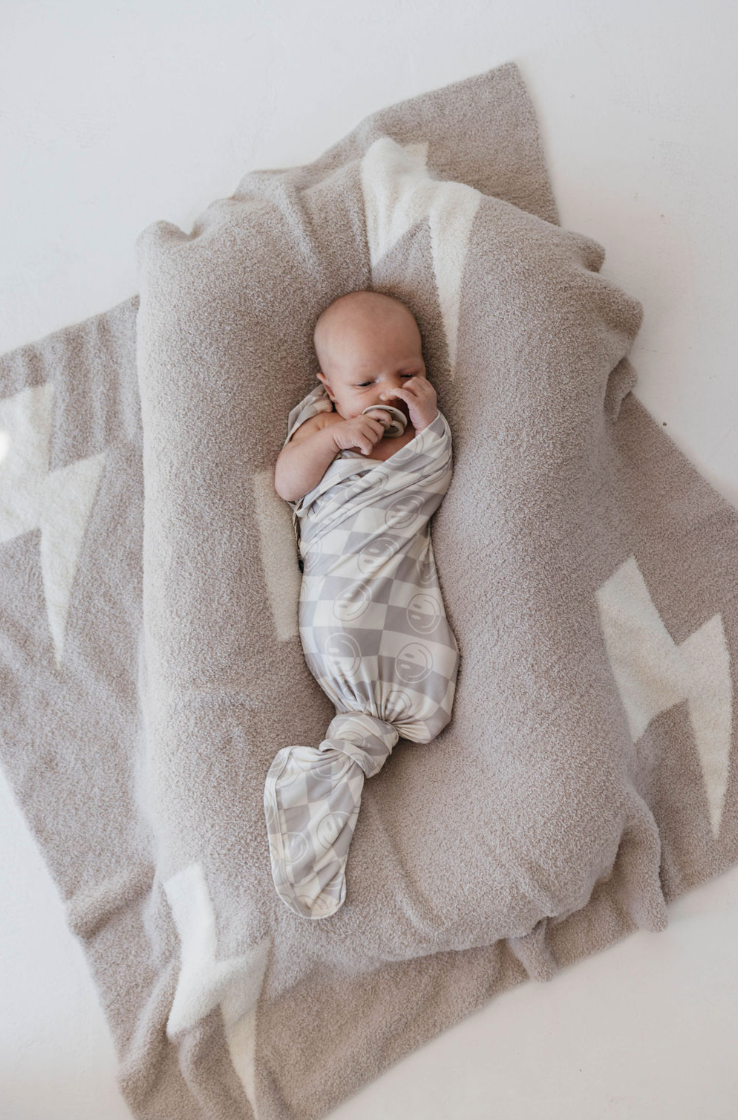 An infant is cozily wrapped in the Bamboo Swaddle | Smile Checkerboard by forever french baby, resting on a soft, cushioned mat. The mat is set atop a larger beige blanket adorned with white zigzag patterns. The baby seems to be sleeping peacefully with one hand near their mouth.