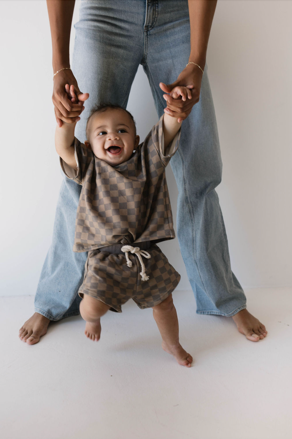 A smiling baby dressed in a Short Set | Faded Brown Checkerboard from forever french baby's trendy kids fashion collection is held up by an adult wearing blue jeans. The baby's hands are securely gripped by the adult as their tiny feet lift off the ground. The plain white background ensures all attention is on this joyous moment.