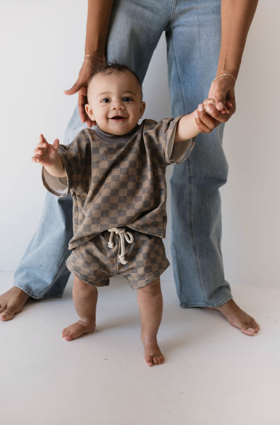 A baby dressed in the stylish "Short Set | Faded Brown Checkerboard" from forever french baby stands on a white floor, holding an adult's hands for support. Both are barefoot, with the adult wearing blue jeans. The baby gleefully smiles at the camera, highlighting adorable kids' fashion.