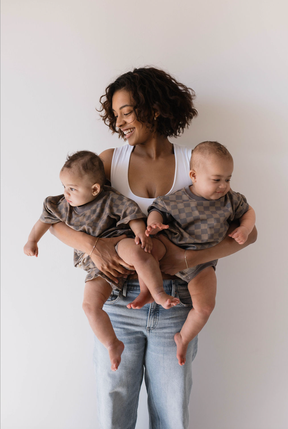 A smiling woman with curly hair, wearing a white top and jeans, holds two babies dressed in matching Short Sets | Faded Brown Checkerboard by forever french baby. The kids' fashion is on point as the babies, held one on each arm, gaze curiously in different directions against the plain white background.