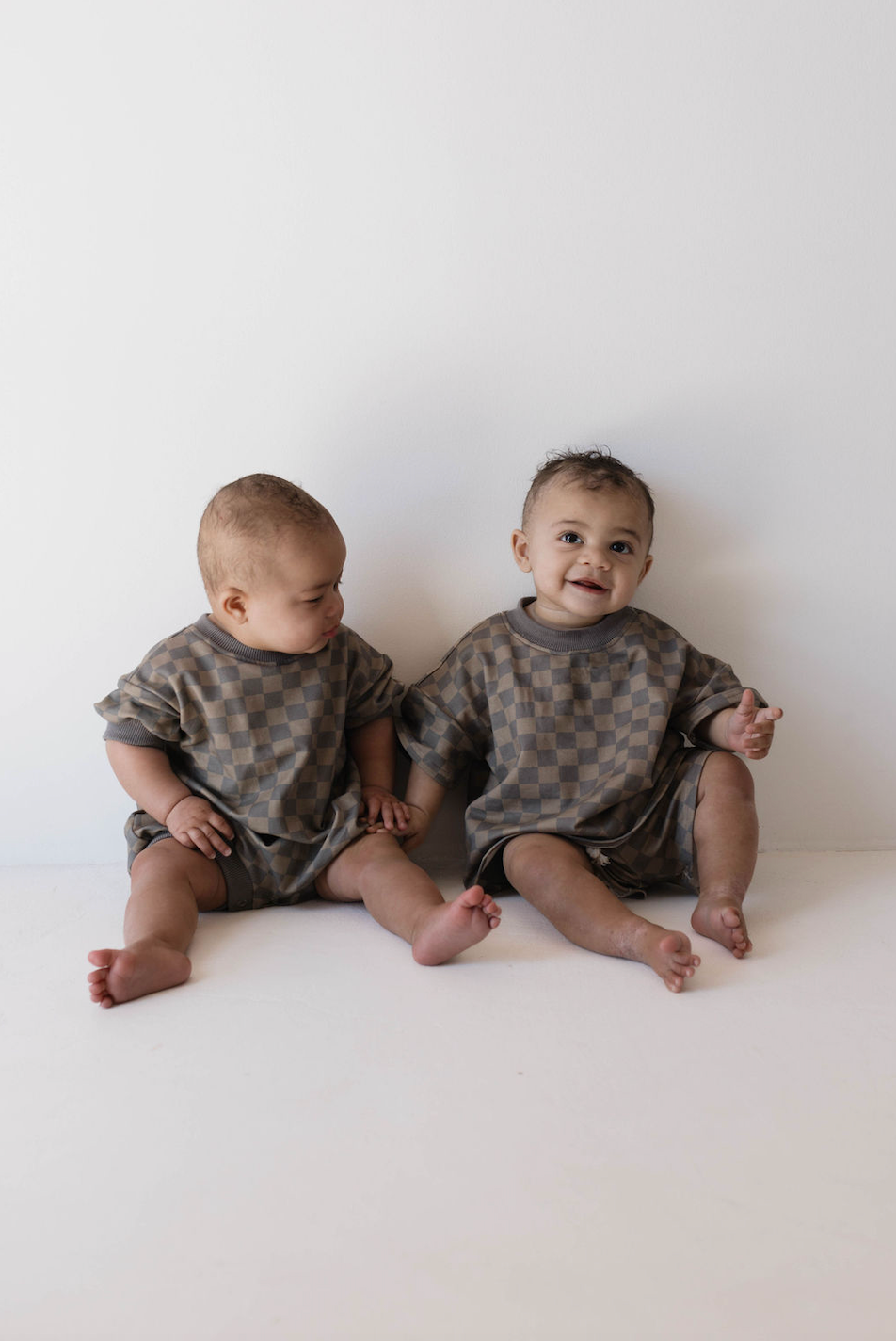 Two babies sit side by side on the floor against a white background, donning adorable Short Sets in Faded Brown Checkerboard. The baby on the left looks down at their hands while the baby on the right looks up and smiles at the camera, showcasing their charming kids' fashion from forever french baby.