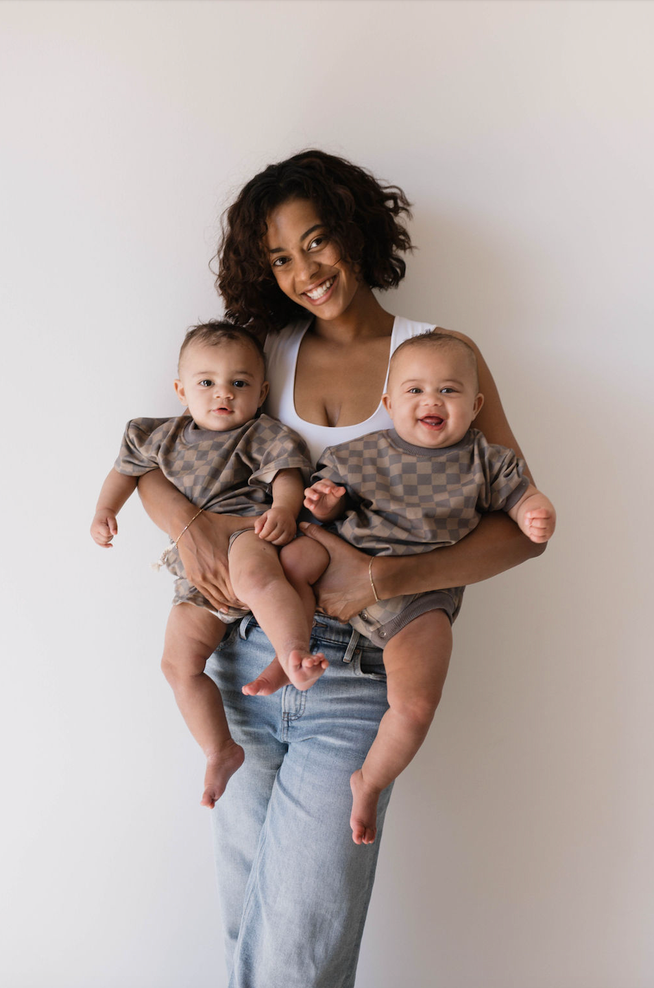 A smiling woman with curly hair, dressed in a white tank top and jeans, is holding two babies. The babies are styled in the latest kids' fashion from forever french baby, wearing matching Short Sets in Faded Brown Checkerboard. Each baby is positioned on one of her arms, with one showing a neutral expression and the other laughing joyfully.