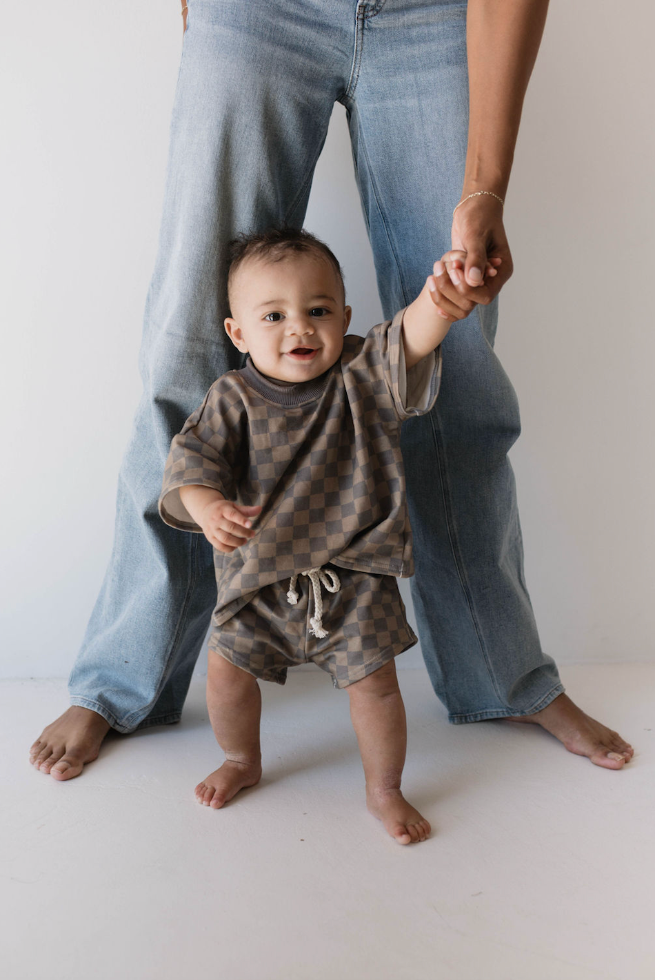 A baby in a "Short Set | Faded Brown Checkerboard" by forever french baby holds onto an adult's hands for support. The adult, dressed in blue jeans, stands behind the child. The baby smiles and appears to be learning to walk in a well-lit room with a white background, showcasing the latest in kids fashion.