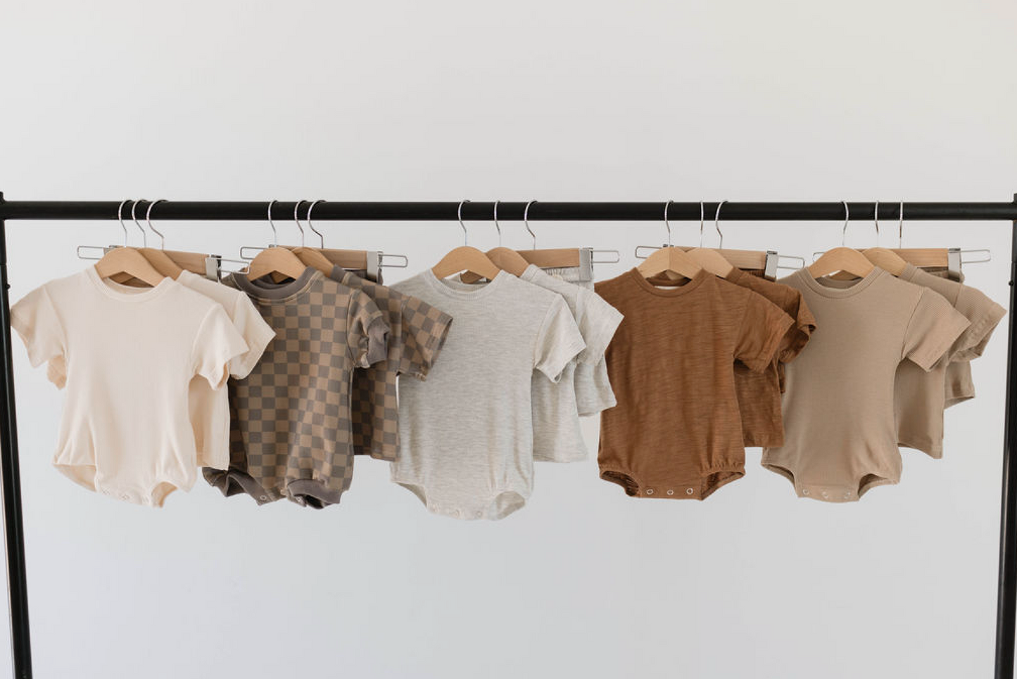 A row of baby onesies in various neutral colors hang on wooden hangers from a black clothes rack against a white background. The kids' fashion display includes solid colors and the "Short Set | Faded Brown Checkerboard" from forever french baby, showcasing adorable options for little ones.