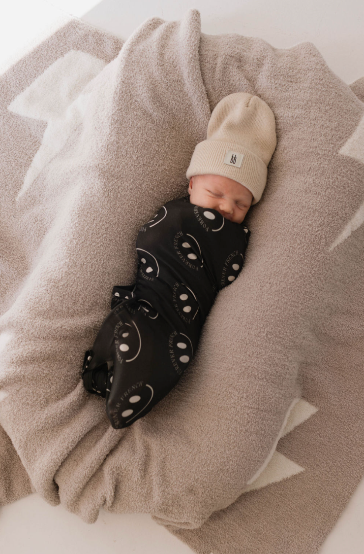 A newborn baby is swaddled in the Bamboo Swaddle | Charcoal & White ff Smile from forever french baby, and wearing a beige knit hat. The baby is lying on a soft, light-colored blanket with white star designs, placed on a white surface. The baby appears to be sleeping peacefully.