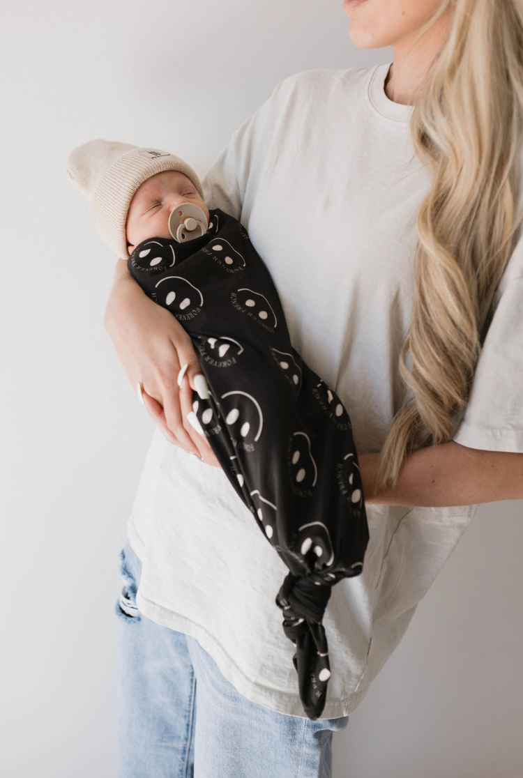 A person wearing a white t-shirt and blue jeans cradles a sleeping baby wrapped in the "Bamboo Swaddle | Charcoal & White ff Smile" by forever french baby. The baby, covered in hypo-allergenic clothing and sporting a beige knit hat, peacefully sucks on a pacifier. The adult has long, wavy blonde hair, and the background is plain white.