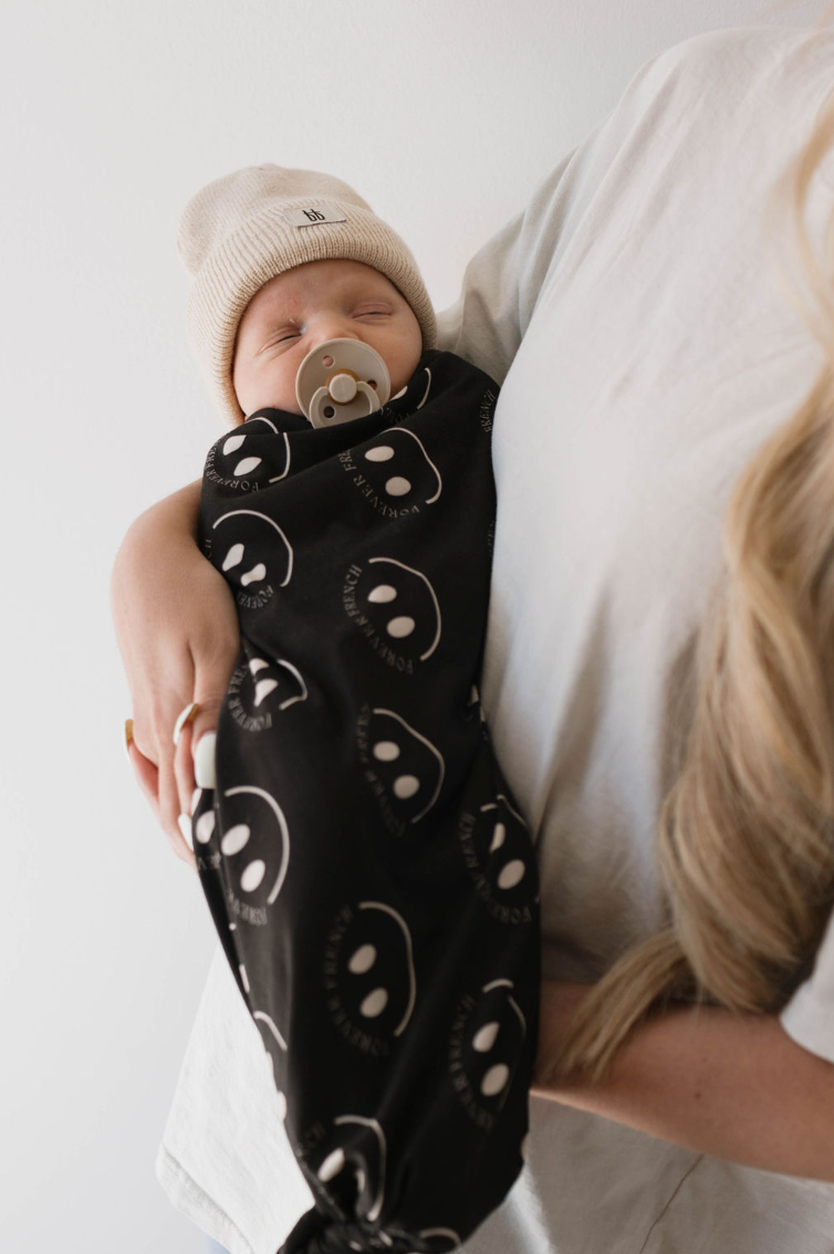 A baby wearing a beige knit hat and a pacifier is swaddled in the "Bamboo Swaddle | Charcoal & White ff Smile" by forever french baby. The baby is being held by an adult with long blonde hair, whose face is not visible but is wearing a light-colored shirt.