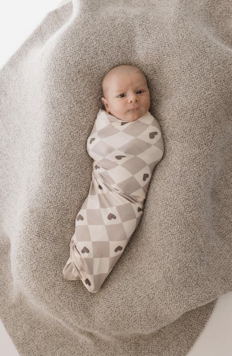 A baby wrapped in a Bamboo Swaddle | Check your Heart from forever french baby is lying on a soft, textured blanket. The baby has a thoughtful expression, and the overall scene is cozy and neutral-toned.