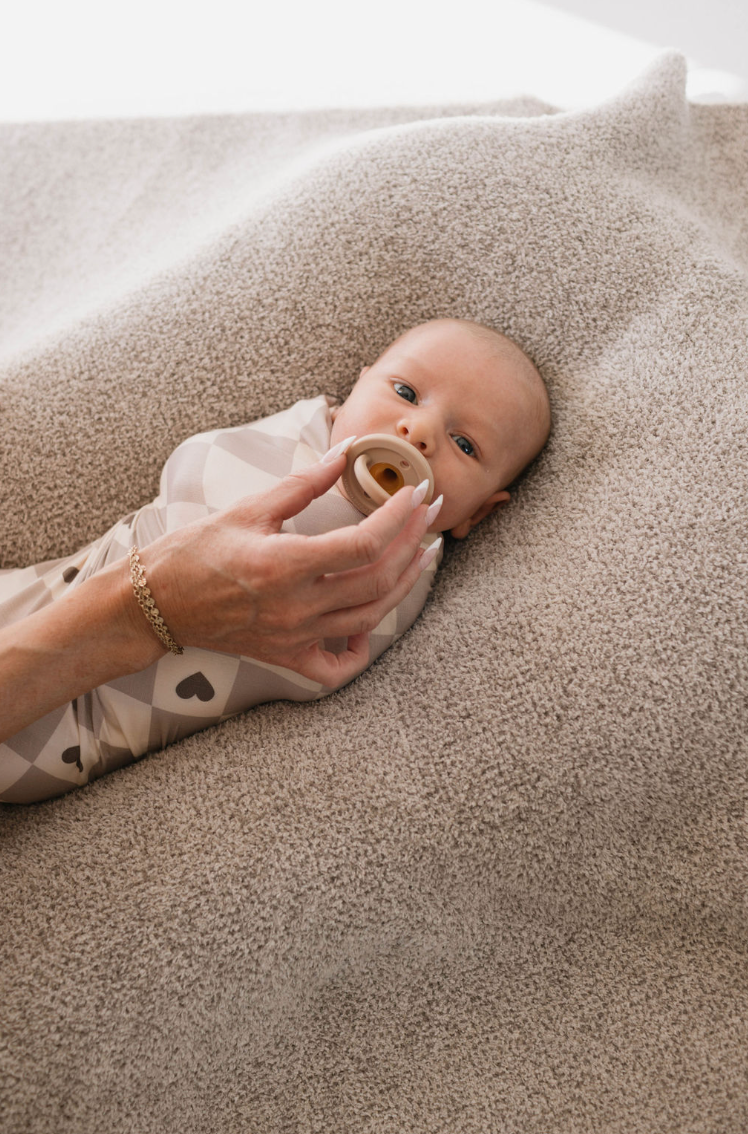 A baby is lying on a soft, beige blanket, wrapped snugly in a "Check your Heart" Bamboo Swaddle by forever french baby. An adult hand is gently holding a pacifier near the baby's mouth. The baby has a calm expression and is gazing upward, comfortably nestled in breathable baby sleepwear.