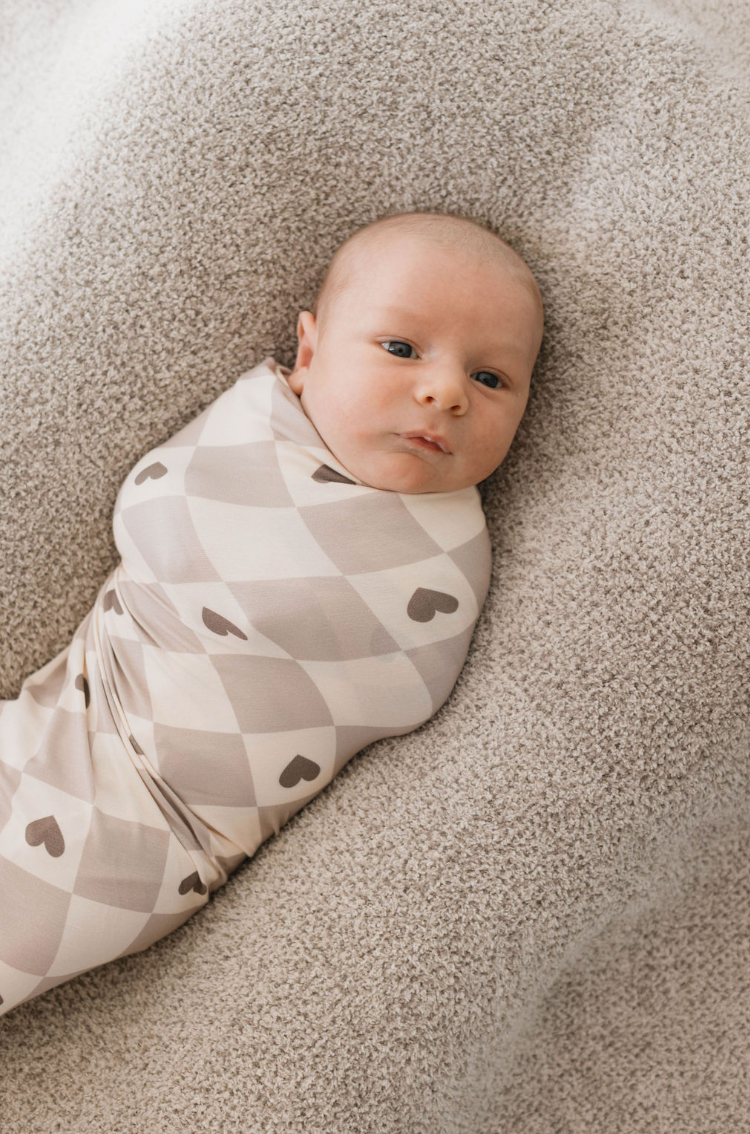 A baby lies swaddled in a soft, "Bamboo Swaddle | Check your Heart" blanket from forever french baby, adorned with small heart shapes. The baby, with eyes open and a calm expression, is resting on a textured, beige surface.