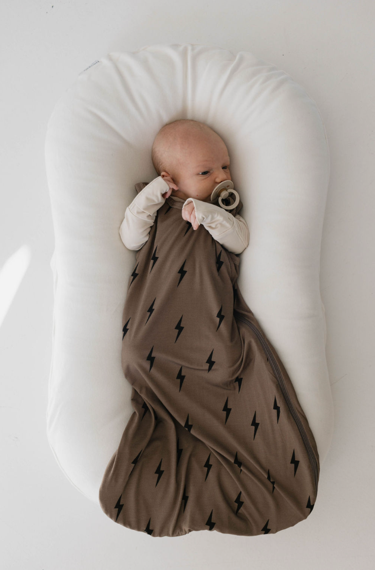 A baby sleeps calmly in a cushioned, white baby lounger. The baby is swaddled in a forever french baby's Bamboo Sleep Sack in brown with black lightning bolt patterns and has a pacifier in its mouth. The background is plain white, highlighting the peaceful setting and ensuring all of the baby's sleep needs are met.