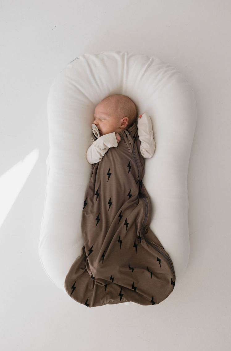 A sleeping baby lies in a white, cushioned baby lounger. The baby is snuggled in the Forever French Baby Bamboo Sleep Sack with brown and black lightning bolt patterns and has a pacifier in their mouth. The background is plain white, ensuring the baby's sleep needs are met in the softest, most comfortable way.