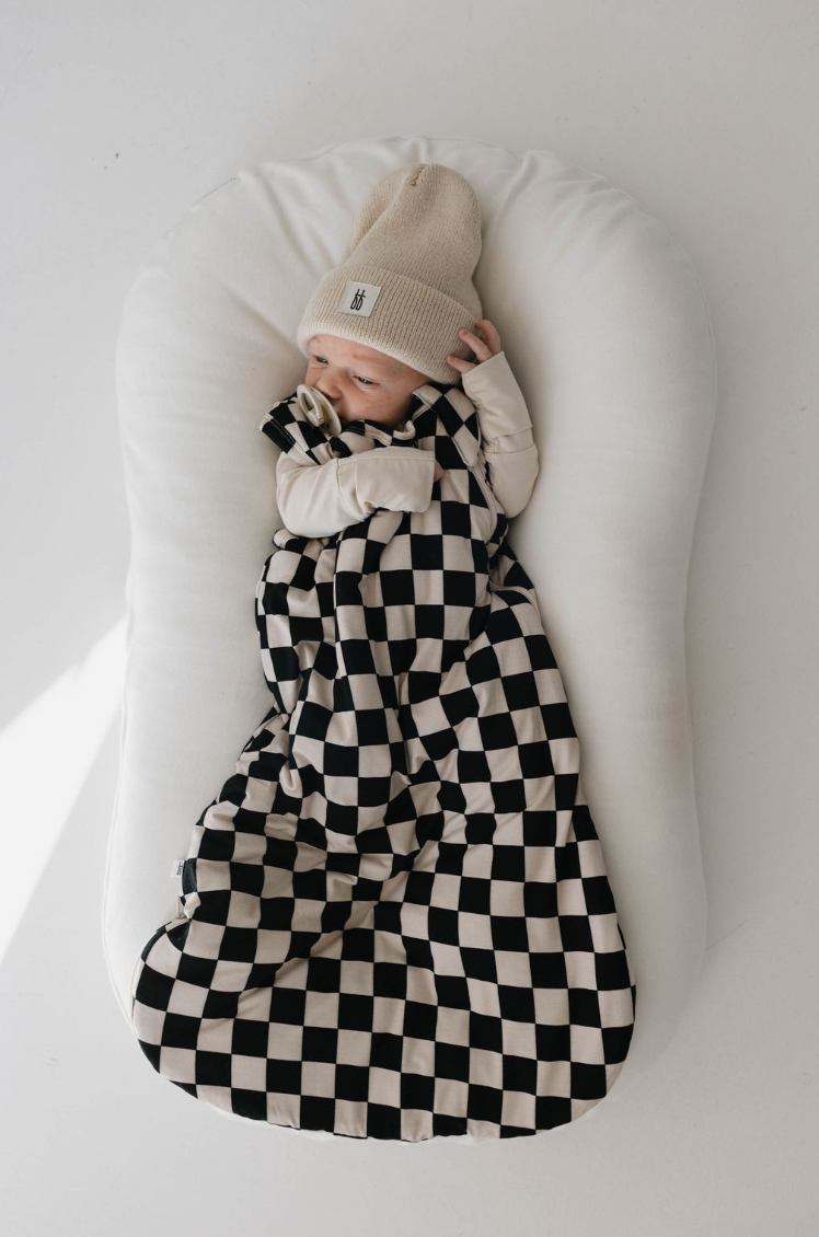 A baby, wearing a beige hat, lies in a cushioned baby lounger wrapped in the Bamboo Sleep Sack in Black Checkerboard by Forever French Baby. With one hand near its mouth and looking to the side, the baby is snugly meeting all of its sleep needs.