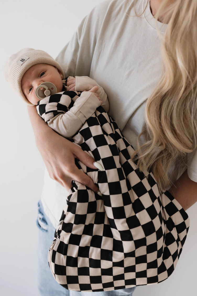 A person with long wavy blonde hair, wearing a light t-shirt, holds a baby wrapped in the Bamboo Sleep Sack | Black Checkerboard by forever french baby. The baby is wearing a cream-colored beanie and has a pacifier in their mouth.