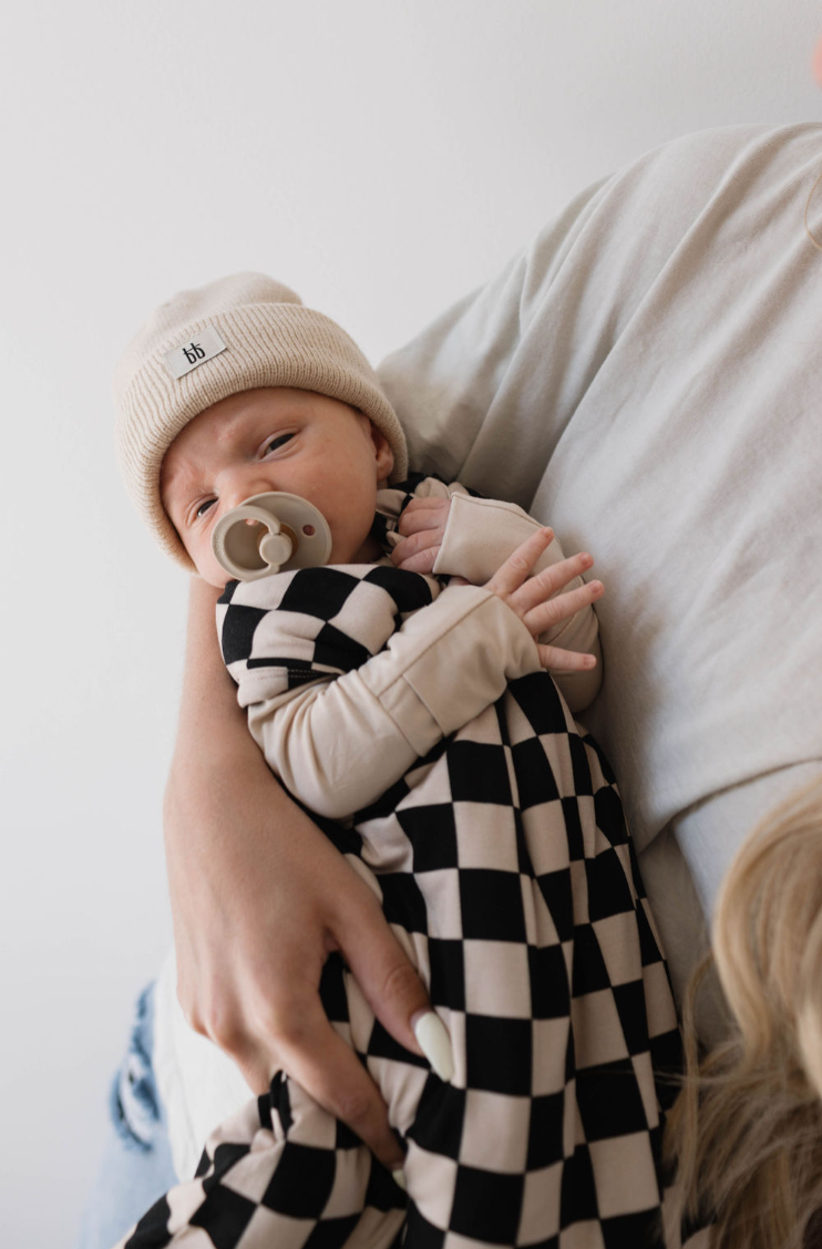 A baby wearing a knit hat and pacifier is cradled in an adult's arms. The baby, dressed in the forever french baby Bamboo Sleep Sack | Black Checkerboard, is nestled comfortably. The adult, visible from the shoulder and wearing a light-colored top, watches over as the baby's eyes are partially closed.