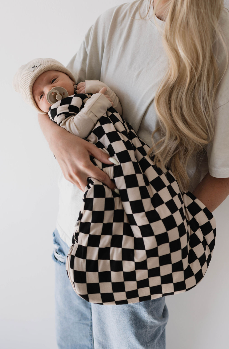 A person with long, blonde hair holds a baby wrapped in the forever french baby Bamboo Sleep Sack in Black Checkerboard. The baby is wearing a beige hat and has a pacifier in their mouth. Both the person and the baby are dressed in light-colored clothing, ideal for meeting the baby's sleep needs.