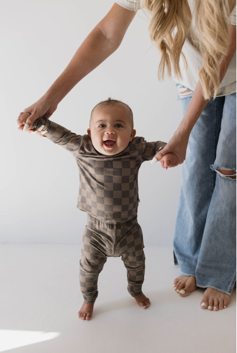 A smiling baby in the Forever French "Sleepy Time Set" with a faded brown checkerboard pattern, made from breathable bamboo fabric, is held up by an adult's hands. The adult, dressed in a white shirt and ripped blue jeans, is assisting the baby in standing. The background is a plain white wall, and both are barefoot.