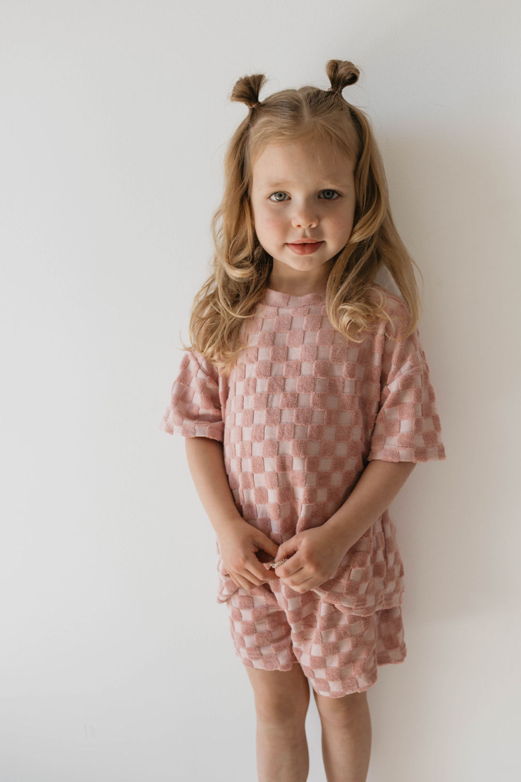 A young girl with light brown hair styled in two small buns stands against a plain white background. She wears the forever french baby Terry Checkerboard Short Set in Perfect Pink, featuring a short-sleeved shirt and matching shorts from her summer wardrobe. A slight smile graces her face.