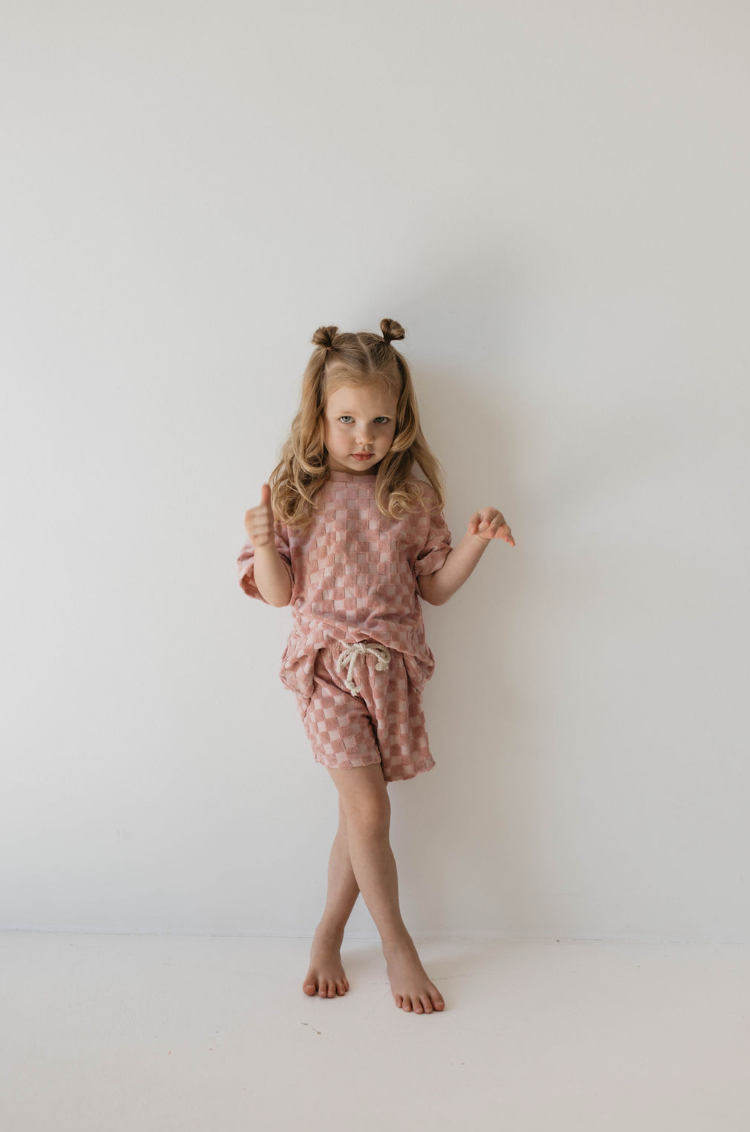 A young girl with long, wavy hair styled in two pigtail buns stands against a plain white wall. She wears the forever french baby Terry Checkerboard Short Set in Perfect Pink, adding a fun touch to her summer wardrobe. Barefoot and playful, she holds her hands up in a gesture, gazing slightly to the side.