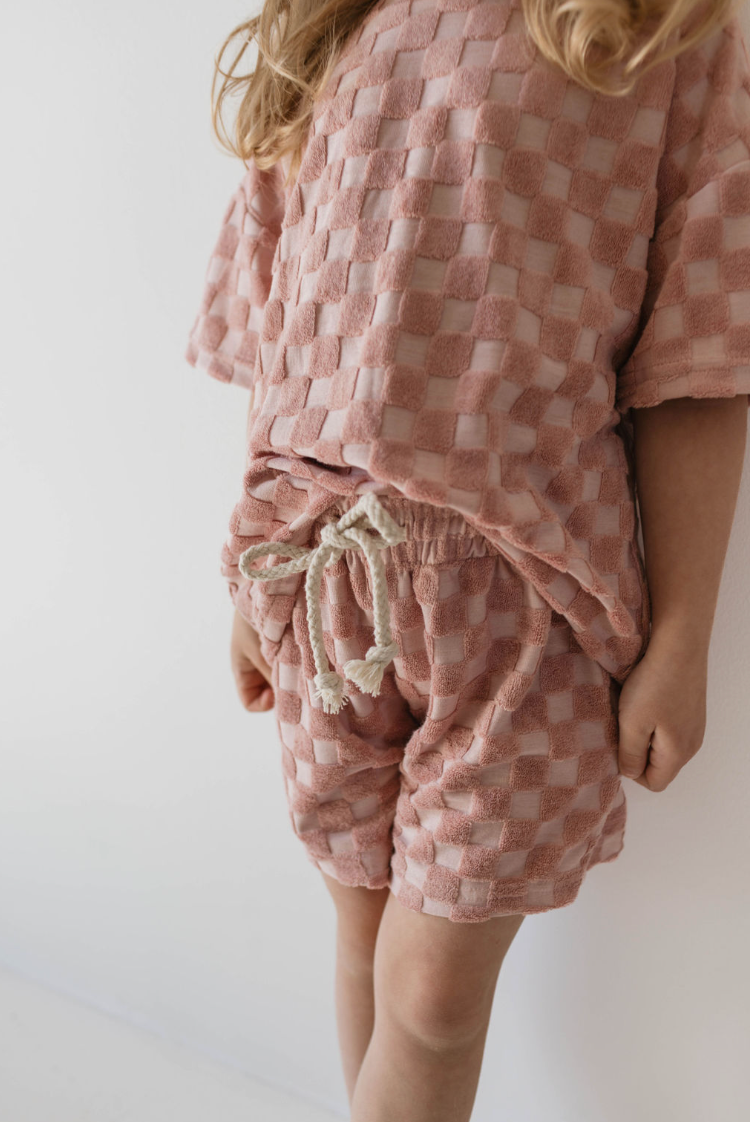 A child wearing the Terry Checkerboard Short Set in Perfect Pink from forever french baby stands against a white backdrop. This terry cloth outfit features a textured checkered pattern, with shorts secured by a white drawstring. The child's face is not visible.