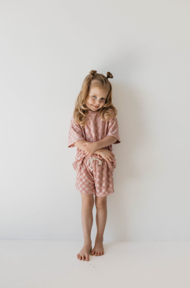 A young girl stands against a plain white background with her long, wavy hair styled in two small buns on top. She is wearing the Terry Checkerboard Short Set in Perfect Pink by forever french baby, featuring a loose t-shirt and matching shorts. With her hands clasped in front and barefoot, she embodies the ideal summer wardrobe.