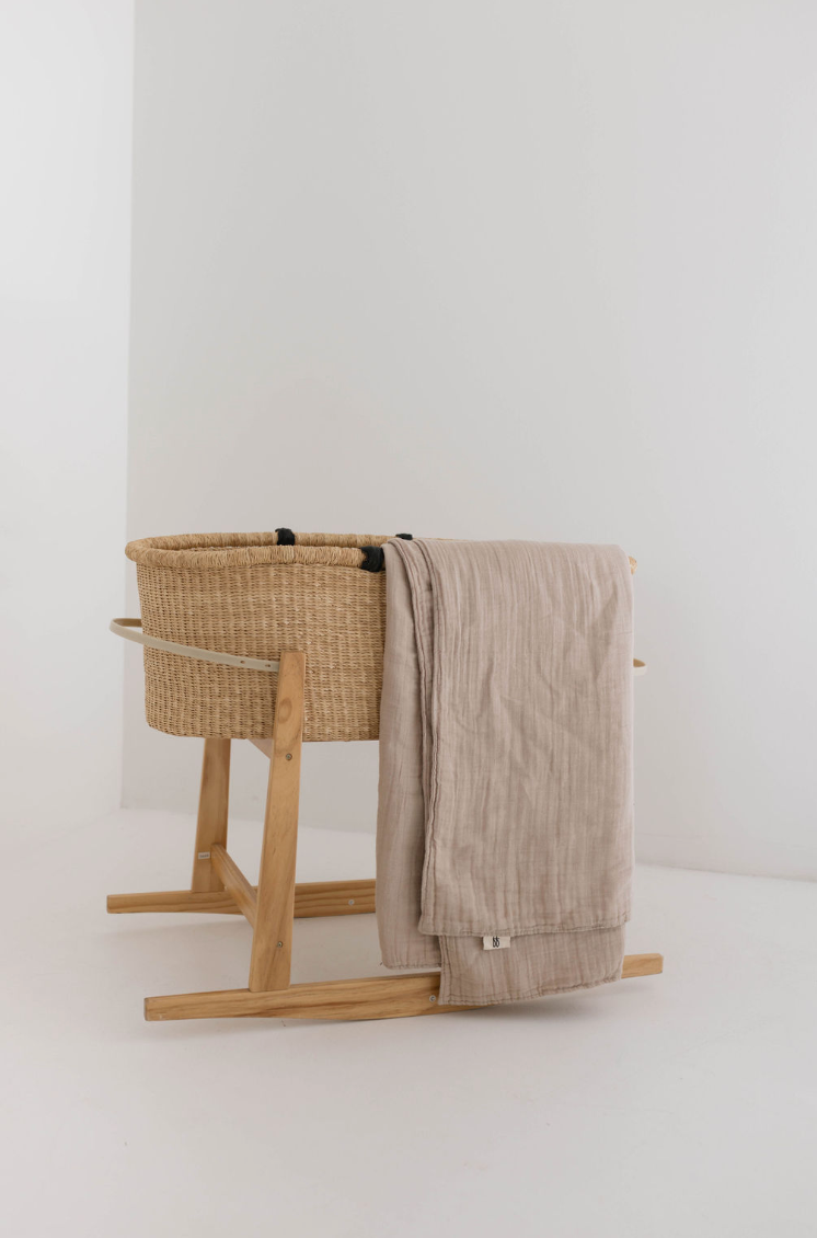 A minimalist baby bassinet made of woven natural fibers on a wooden stand, featuring the high-quality Muslin Throw | Natural by forever french baby draped over the side. The light beige, textured fabric complements the modern decor, all set against a plain white background.