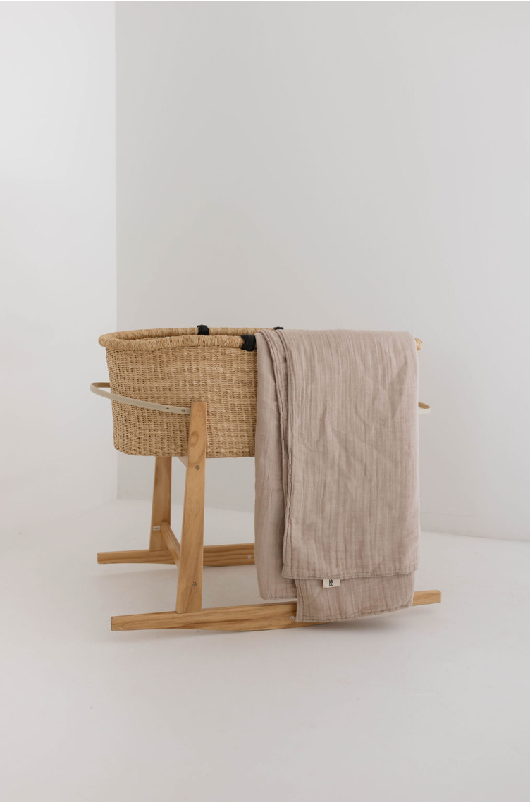 A wicker baby bassinet on a wooden stand, placed in a minimalist white room. A Muslin Throw | Natural from forever french baby drapes elegantly over the side of the bassinet. The modern decor’s overall aesthetic is simple and clean.