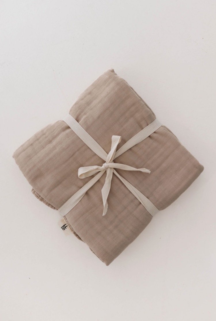 The Muslin Throw | Natural by forever french baby is neatly tied with a white fabric ribbon against a white background. The high-quality muslin, in a soft beige hue, has a textured, slightly crinkled appearance that offers a cozy look, perfectly complementing modern decor.