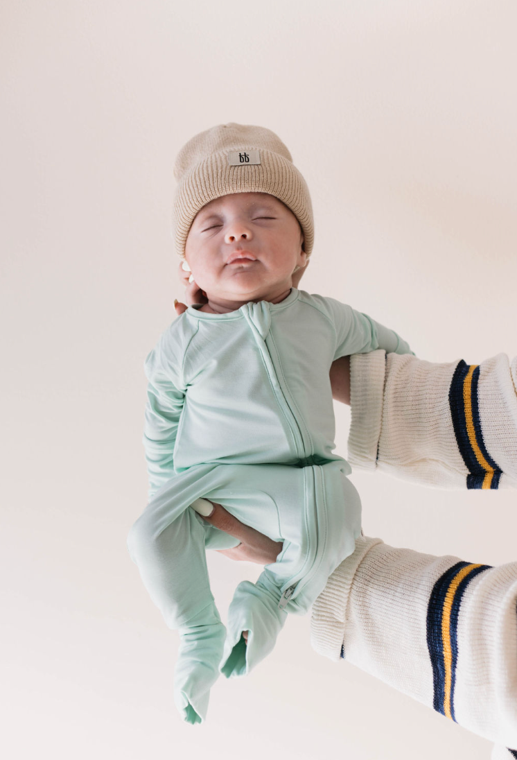 A baby in forever french baby's Bamboo Zip Pajamas in Mint, a beige knit hat, and a light green onesie is gently held by an adult wearing a white sweater with dark blue and yellow striped sleeves. The baby looks calm and relaxed.