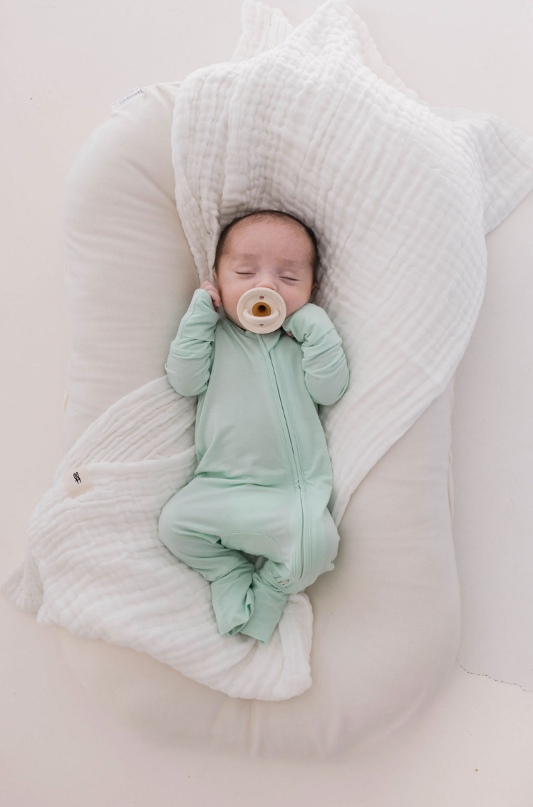 A sleeping baby in mint Bamboo Zip Pajamas by forever french baby rests on a white cushion. With a pacifier and swaddled in a soft, breathable blanket, the scene is calm and serene.